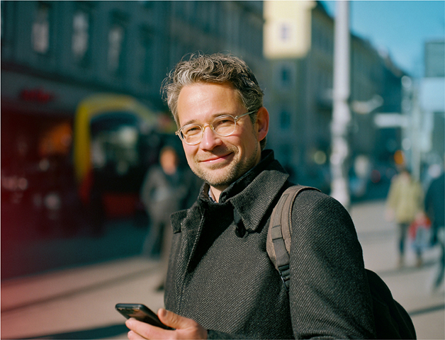 junger Mann im Mantel mit Smartphone in der Hand steht in der Stadt und lächelt in die Kamera