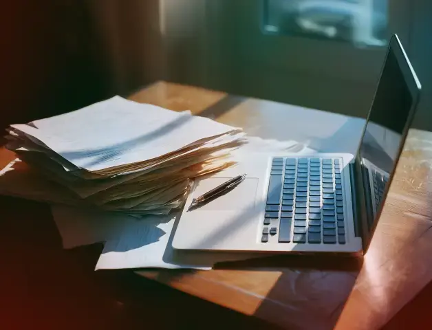 auf einem Tisch steht ein Laptop, davor viele Akten mit Papieren